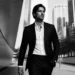 Man in a black suit and white shirt walking in an urban cityscape in black and white.
