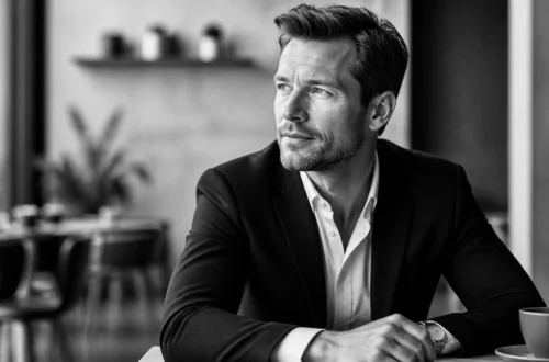 Man in a dark blazer and white shirt sits at a cafe table, looking to the right, with a cup on the table.