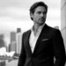 Man in a dark suit and white shirt stands outdoors in an urban setting, looking off to the side.