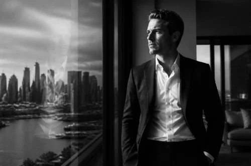 Man in a dark suit gazes out a large window, city skyline reflection visible on the glass.