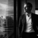 Man in a dark suit gazes out a large window, city skyline reflection visible on the glass.