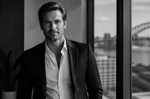 Black-and-white portrait of a man in a suit standing by a window, with an urban skyline and a bridge visible outside.
