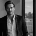 Black-and-white portrait of a man in a suit standing by a window, with an urban skyline and a bridge visible outside.