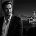 Man in a dark suit leaning on a railing at night, with a lit city skyline in the background.