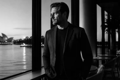 Man in a dark blazer leaning on a railing beside the water, looking pensively to the left with a cityscape and the Sydney Opera House in the distance (black-and-white).