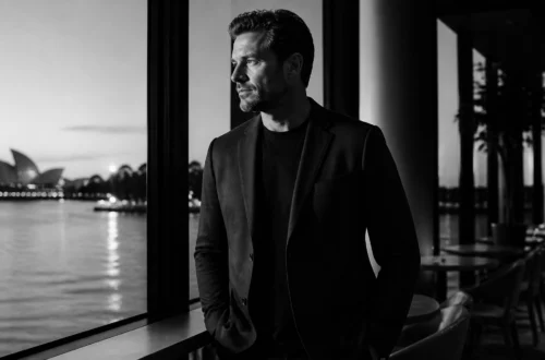 Man in a dark blazer leaning on a railing beside the water, looking pensively to the left with a cityscape and the Sydney Opera House in the distance (black-and-white).