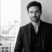 Professional portrait of a man in a black suit posing in a modern office with a city skyline in the background (black & white)